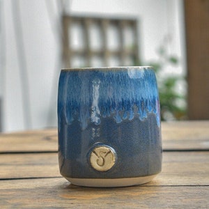 May include: A blue and white ceramic mug with a white glaze and a small, white, circular design on the bottom. The mug is sitting on a wooden table.