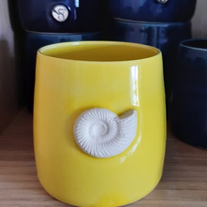 May include: A bright yellow ceramic mug with a white ceramic ammonite shell design.