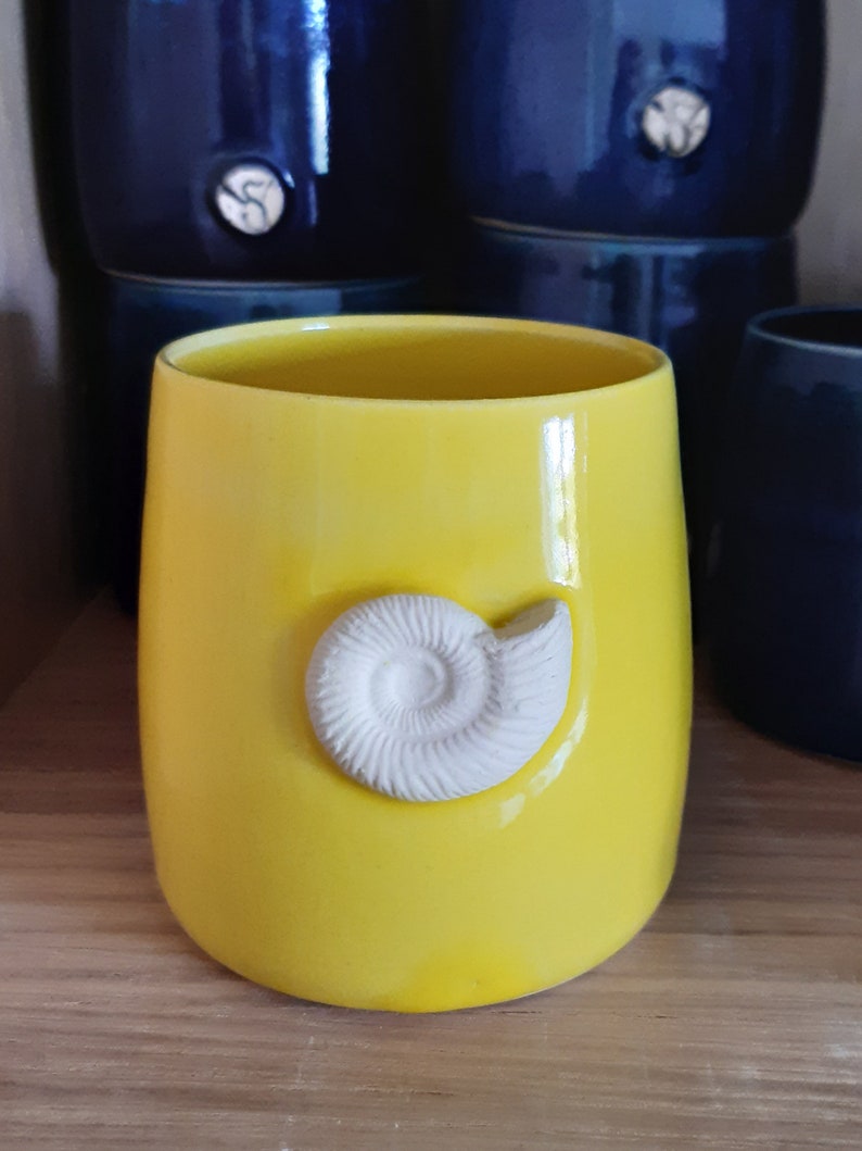 May include: A bright yellow ceramic mug with a white ceramic ammonite shell design.