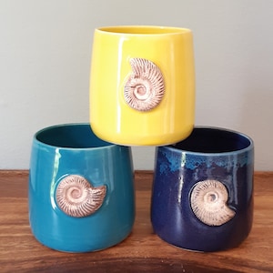 May include: Three ceramic mugs with a unique design. Each mug features a different colour: yellow, teal, and dark blue. Each mug has a raised, brown ammonite fossil design.