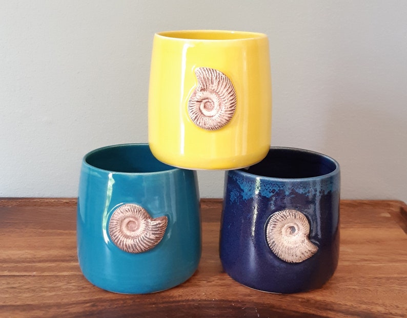 May include: Three ceramic mugs with a unique design. Each mug features a different color: yellow, teal, and dark blue. Each mug has a raised, brown ammonite fossil design.