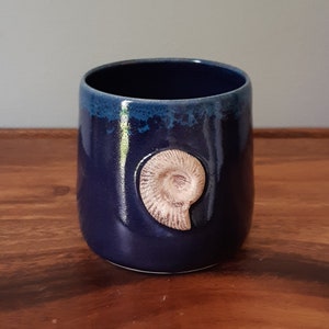 May include: A dark blue ceramic mug with a light brown ammonite fossil design. The mug has a textured glaze.