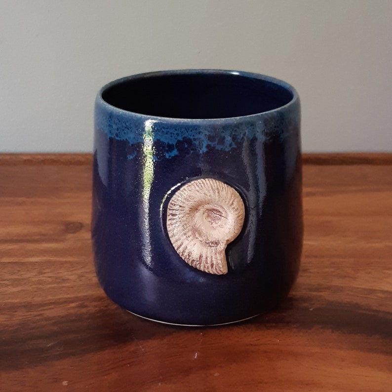 May include: A dark blue ceramic mug with a light brown ammonite fossil design. The mug has a textured glaze.