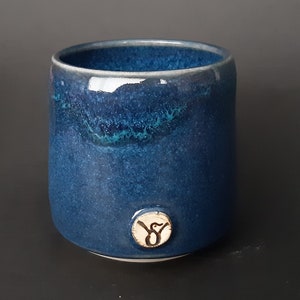 May include: A deep blue ceramic mug with a white glaze and a small, white, circular logo featuring a bird design. The mug has a speckled, textured surface.
