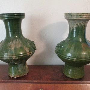 May include: Two green ceramic vases with a rounded body and a narrow neck. The vases have a textured surface and are decorated with raised details.