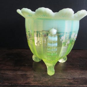 May include: A vintage, light green glass bowl with a ruffled rim and three feet. The bowl has a ribbed texture with floral and leaf designs. The rim and handle have a lighter, almost white, color.