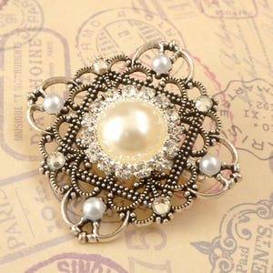 May include: A silver-toned brooch with a large faux pearl surrounded by rhinestones and smaller faux pearls. The brooch has a delicate, intricate design.