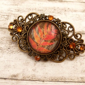 May include: A bronze hair clip with an ornate design features a round glass cabochon with a red and orange leaf pattern. The clip is decorated with small orange rhinestones.
