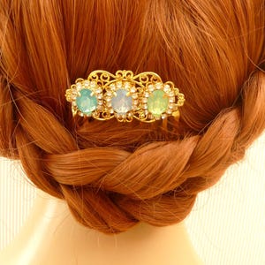 Könnte beinhalten: Ein goldener Haarkamm mit drei ovalen blauen und grünen Edelsteinen. Der Kamm befindet sich im Haar einer Person mit langen, roten Haaren.