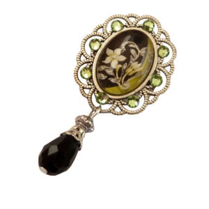 May include: A silver pendant with a black and green floral design in a cameo setting. The pendant has a black teardrop-shaped bead hanging from it.