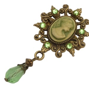 Op de afbeelding: Een gouden hanger met een cameo van een vrouwenhoofd omringd door groene strass steentjes. De hanger is bevestigd aan een groene kristallen druppel.
