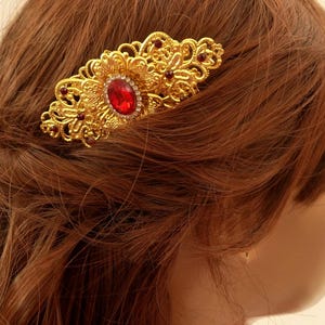 May include: A gold-colored hair clip with an ornate, filigree design. The clip features a large, oval red gemstone in the center, surrounded by smaller red accents and clear crystals. The clip is set against auburn hair.