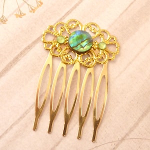 Puede incluir: Un peine para el cabello dorado con un diseño floral decorativo. El peine presenta una concha de abulón ovalada central y dos piedras verdes más pequeñas. Las púas del peine son largas y delgadas, creando un accesorio elegante.