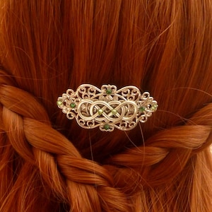May include: A silver hair clip with a Celtic knot design and green gemstones. The clip is in the shape of an oval and has a delicate, intricate design.