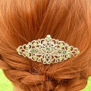 Könnte beinhalten: Silberfarbene Haarklammer mit einem aufwendigen keltischen Knotenmuster, verziert mit kleinen grünen Perlen. Die Spange ist vor dem Hintergrund von geflochtenem, rotbraunem Haar platziert. Ein dekoratives Haaraccessoire.