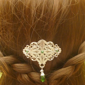 Op de afbeelding: Een zilverkleurige haarclip met Keltische knoop, groene accenten en een bungelende groene druppelvormige bedel. Het sierlijke haaraccessoire is geplaatst tegen een achtergrond van gevlochten bruin haar.