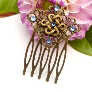 Puede incluir: Un peine para el cabello dorado con un diseño decorativo que presenta un nudo y pedrería azul. El peine tiene cinco dientes.