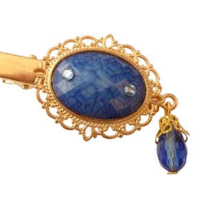 May include: A gold hair clip with a blue and white agate stone in a decorative frame. A dangling blue glass bead hangs from the bottom of the clip.