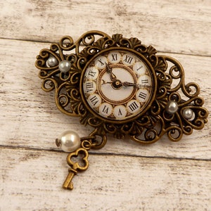 Op de afbeelding: Een gouden metalen broche met een klokgezicht en sierlijke details. Het klokgezicht heeft Romeinse cijfers en een sleutelgatontwerp. De broche is versierd met witte parels.