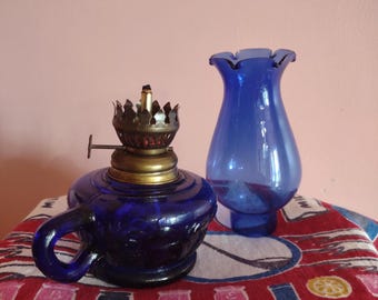 May include: A cobalt blue glass oil lamp base with a handle and a brass burner mechanism, accompanied by a separate matching cobalt blue glass chimney with a ruffled top edge. The lamp features a deep blue color and a textured glass body design.