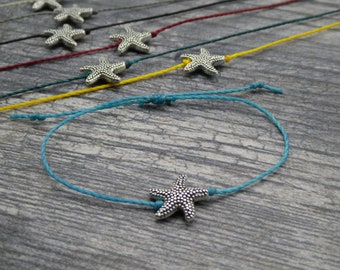 Puede incluir: Un brazalete de cuerda azul turquesa con un dije de estrella de mar plateada. El brazalete está atado con un nudo simple y se muestra sobre un fondo de madera.