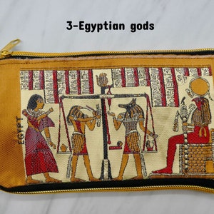 May include: A gold zippered pouch with a beige background and a tapestry design featuring three Egyptian gods. The gods are depicted in a scene with a scale, and the text "EGYPT" is visible on the pouch.