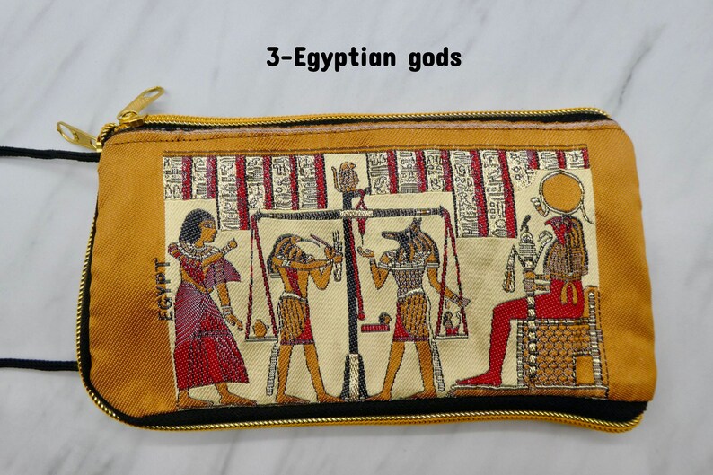 May include: A gold zippered pouch with a beige background and a tapestry design featuring three Egyptian gods. The gods are depicted in a scene with a scale, and the text "EGYPT" is visible on the pouch.
