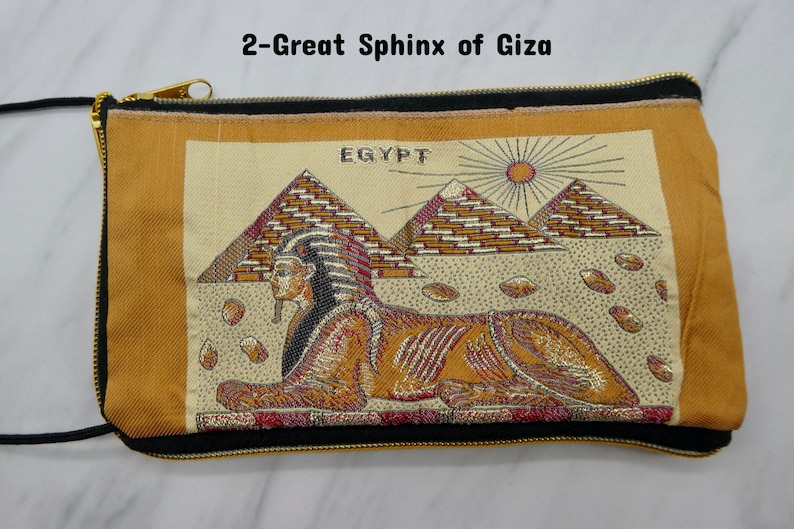 May include: A gold and brown zippered pouch with a black trim. The pouch features an embroidered image of the Great Sphinx of Giza in front of the pyramids. The image also includes the word "EGYPT" and a sun.