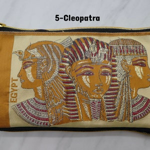 May include: A brown and beige zippered pouch with a black strap. The pouch features an embroidered image of three women with gold and red headdresses. The text "EGYPT" is embroidered vertically on the left side of the pouch. The text "5-Cleopatra" is printed above the pouch.