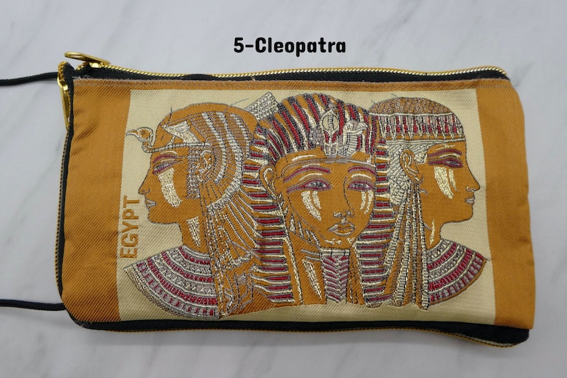 May include: A brown and beige zippered pouch with a black strap. The pouch features an embroidered image of three women with gold and red headdresses. The text "EGYPT" is embroidered vertically on the left side of the pouch. The text "5-Cleopatra" is printed above the pouch.