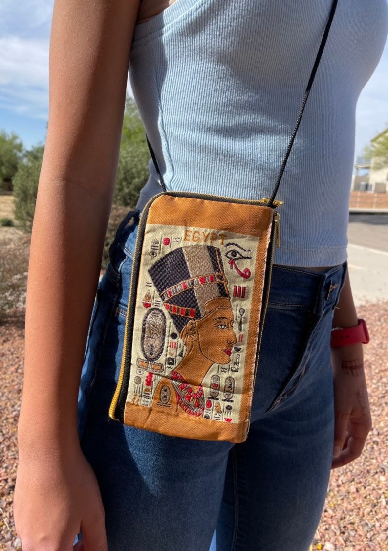 May include: A brown crossbody bag with a zipper closure. The bag features an embroidered image of an Egyptian queen with the word "EGYPT" above her.