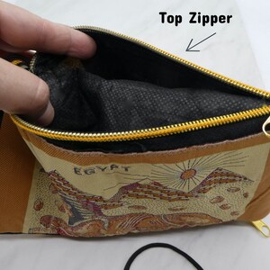 May include: A brown and beige fabric pouch with a black zipper and a gold zipper pull. The pouch has a black fabric lining and the words "EGYPT" printed on the front. The pouch has a black drawstring.