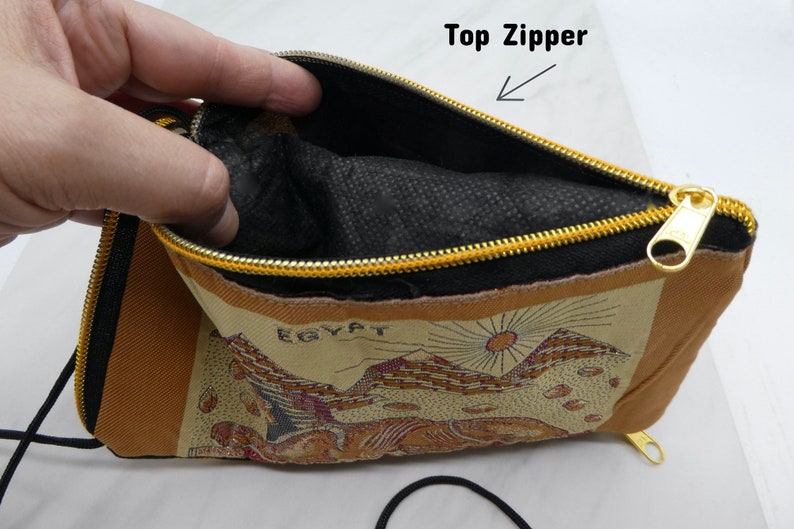 May include: A brown and beige fabric pouch with a black zipper and a gold zipper pull. The pouch has a black fabric lining and the words "EGYPT" printed on the front. The pouch has a black drawstring.