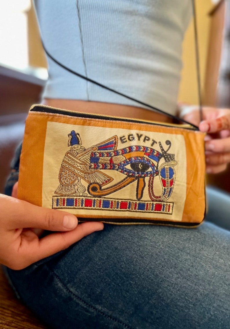 May include: A small, tan crossbody bag with a zipper closure. The bag features an embroidered design of the Eye of Horus with the word "EGYPT" above it.