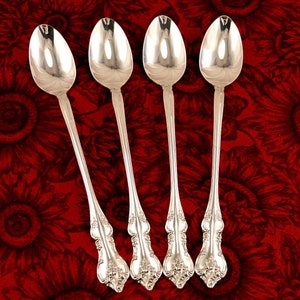 May include: Four silver teaspoons with ornate handles. The spoons are arranged in a row on a red background with a floral pattern.