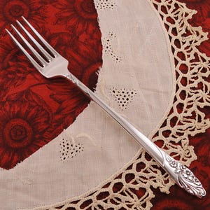 May include: A silver fork with a floral design on the handle. The fork is lying on a white lace tablecloth with a red floral pattern underneath.