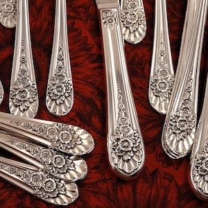 May include: A set of silver-plated cutlery with ornate floral designs. The handles are decorated with raised flowers and leaves. The cutlery is arranged in a fan shape on a red background.