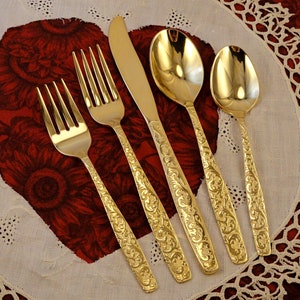 May include: A set of gold-colored flatware, including forks, a knife, and spoons, arranged on a white and red floral patterned surface. The utensils have ornate, engraved handles.