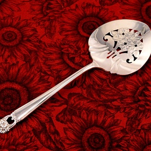 May include: A silver serving spoon with a floral design. The spoon features a decorative floral motif at the bowl and handle end. The spoon is set against a red background with a sunflower pattern.