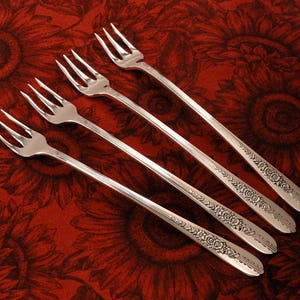 May include: Four silver forks with ornate floral designs on the handles. The forks are arranged in a row, with the tips pointing to the right.