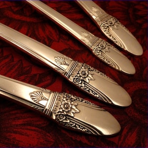 May include: Four silver-plated dinner forks with ornate floral designs on the handles. The forks are arranged in a fan shape on a red fabric background.