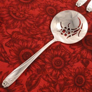 May include: A silver serving spoon with a decorative floral design cut into the bowl. The handle has a floral motif at the end. The spoon is set against a red background with a sunflower pattern.