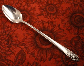 May include: A silver spoon with a floral design on the handle, resting on a red background with a sunflower pattern. The spoon is highly reflective, showing the surrounding environment. The spoon is a piece of silverware.