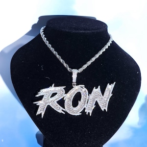 以下が含まれることがあります： キラキラとした透明なクリスタルで「RON」と書かれたシルバーカラーのペンダントネックレス。ペンダントは、お揃いのシルバーロープチェーンから吊り下げられています。ネックレスは黒いバストに飾られています。