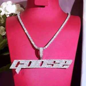 Puede incluir: Un collar plateado con un colgante que deletrea "GOOSE" en una fuente estilizada. El collar y el colgante están incrustados con pequeñas piedras brillantes, probablemente pedrería o cristales. El collar se exhibe en un maniquí rojo.