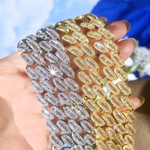 May include: Close-up of two diamond-studded Cuban link chains, one silver and one gold. The chains are made of rectangular-cut stones set in a repeating pattern. The jewelry is displayed against a soft, blurred background.