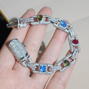 May include: A silver bracelet with a chain link design, encrusted with small, clear crystals. The bracelet features rectangular links with colorful gemstones: blue, green, and red. The clasp is also covered in crystals.