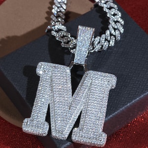 May include: A silver-colored, diamond-encrusted pendant in the shape of the letter "M" hangs from a matching chain. The pendant and chain are set against a dark background, with a hint of red at the bottom. The jewelry sparkles, reflecting light.