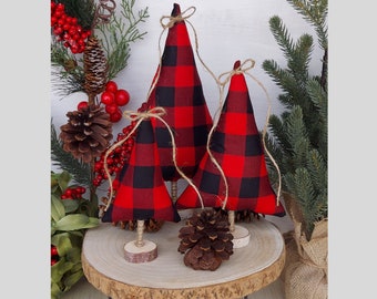 May include: Three decorative Christmas tree ornaments. Each tree is cone-shaped, made of red and black buffalo plaid fabric, and has a twine loop for hanging. The trees are displayed on a wooden slice with a pine cone.
