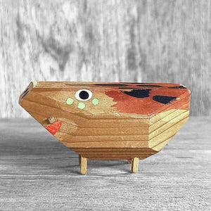 May include: A small wooden sculpture of a fish with a black eye, green spots, and red painted details. The fish is standing on four small legs.